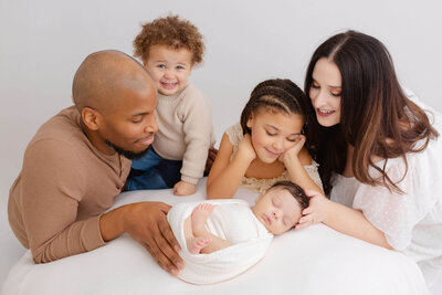 Mom, Dad and two older siblings are sitting next to their newborn baby and looking down on him and smiling. Toddler boy is smiling at the camera. Portland newborn photoshoot