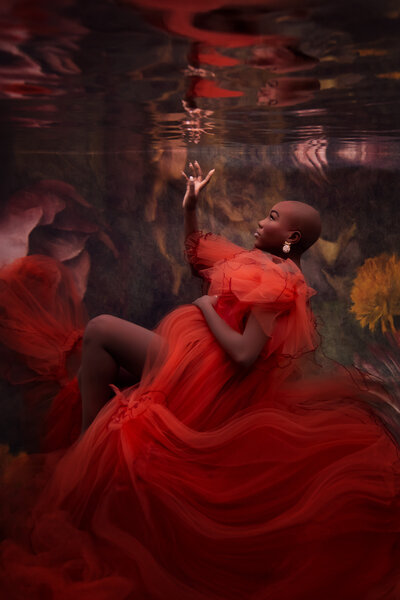 Dark skinned woman in an orange dress for her underwater maternity photoshoot with her hand reaching towards her reflection