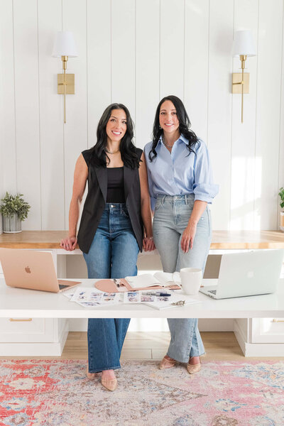 Two women in a modern office; both experts in sales strategy standing behind desk with a laptops, coffee cup, a notebook, and printed papers.