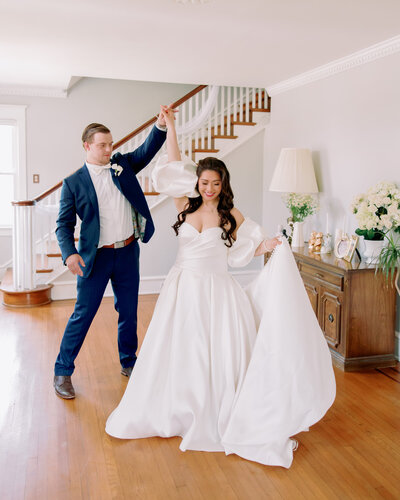 Newlyweds hold hands and dance in a living room.