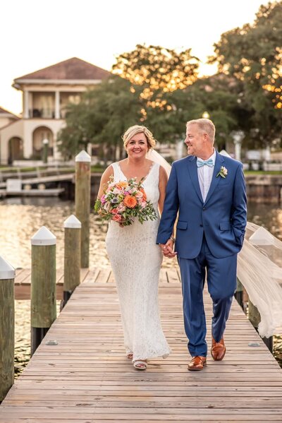 pensacola wedding venues with A bride in a white lace dress holding a colorful bouquet and a groom in a blue suit with a bow tie walk hand in hand on a wooden dock by the water. The background features a serene waterfront with boats, trees, and a house, bathed in warm sunset light at a wedding venue pensaola
