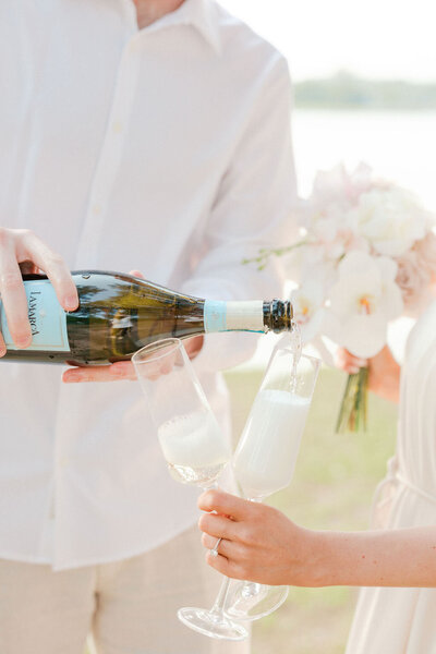 Groom pours champagne bottle into fluted glasses holding bouquet of white flowers by lake Lanier