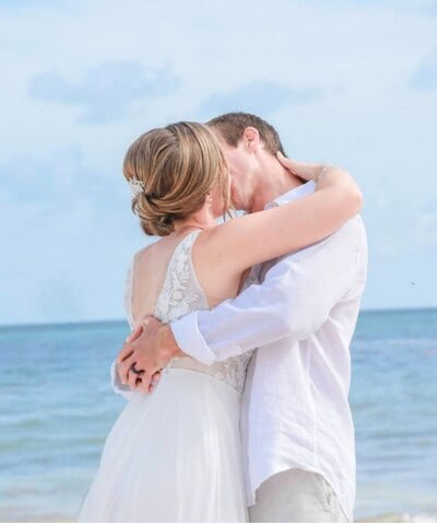 Bride and groom share a kiss on the beach in front of a blue sky and calm ocean during their tropical destination wedding.