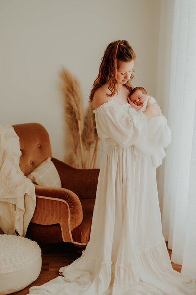 Mother holds her newborn baby to her chest and gazes at the baby. They are standing near a window in  warm  inviting studio