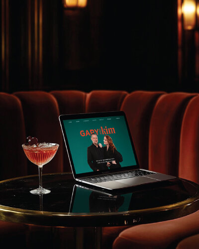 Laptop on a glossy black cocktail lounge table next to an amber drink, displaying the Gary & Kim website—bringing their coaching brand identity to life online.