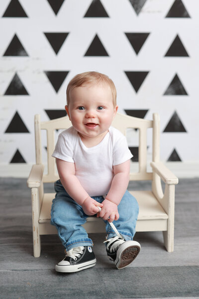 Lehigh Valley Milestone Photographer six month old boy sitting on a small bench wearing a white onesie and jeans with baby Chuck Taylors pulling on the shoelace with a white with black triangles backdrop behind him and a gray wood floor