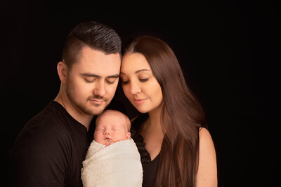 Heartwarming family portrait featuring parents lovingly holding their swaddled newborn against a dark backdrop. Captured in Overland Park, Kansas, this timeless and intimate setup is perfect for families seeking classic and meaningful newborn photography in the Kansas City metro area.