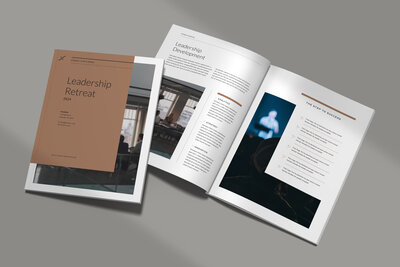 A professional brochure for a leadership retreat displayed on a gray surface. One brochure is closed, showing a minimalist brown and white cover. Another is open, revealing clean layouts with photos of a meeting room and a speaker on stage. The design uses neutral colors, modern typography, and a structured layout aimed at business or corporate audiences.