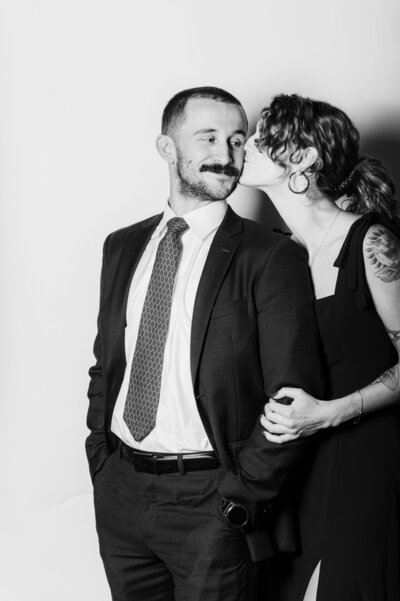 Editorial black and white photo of a couple dressed in formal attire, the woman kissing the man on the cheek, captured against a simple white backdrop.