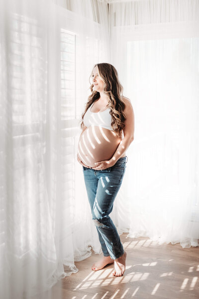 pregnant mother in jeans cradling her belly and looking out the window