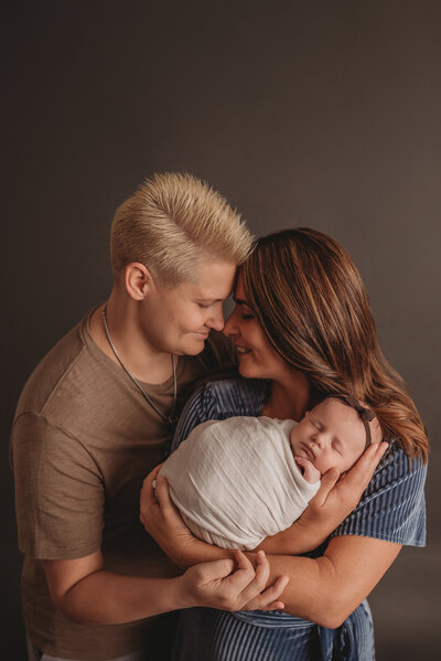 Couple facing head to head smiling holding newborn baby girl asleep and swaddled in white fabric