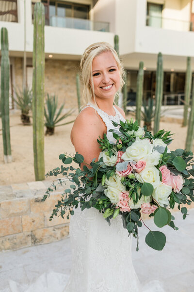 A smiling bride in a lace wedding gown holds a large bouquet of white and pink roses with eucalyptus. She poses outdoors in front of tall cacti at a desert-style venue.