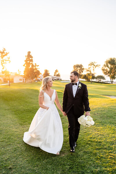 Bride and groom holding hands and walking with the sunset in the background on their wedding day at The La Jolla Country Club wedding venue