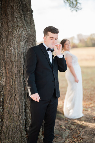 A groom smoking something with a bride standing behind him looking on