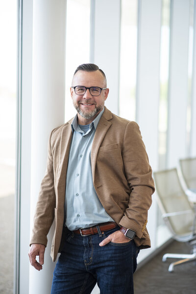 Matthew Kidwell in a brown blazer leaning against window and smiling - Financial Coach at Kidwell Coaching and Consulting 19