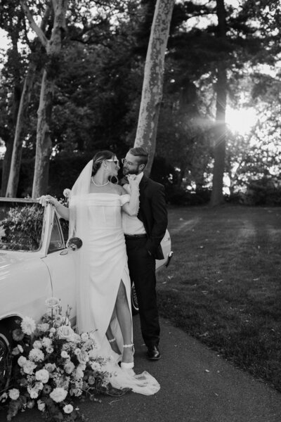 A black and white photo of a bride, wearing a white high slit gown with ankle strapped shoes, and groom, wearing a classic tux with a bow tie, leaning against a classic antique car.