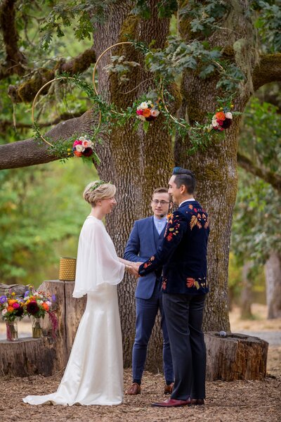A bride and groom stand in front of a tree with a floral design by Flowers and Thyme.