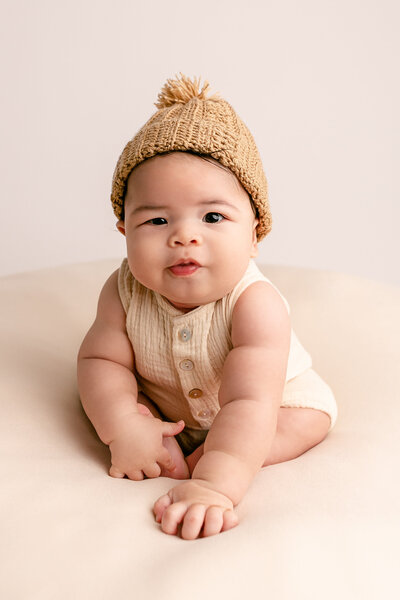 six month old baby in cream romper and brown beanie sitting up and leaning towards camera.