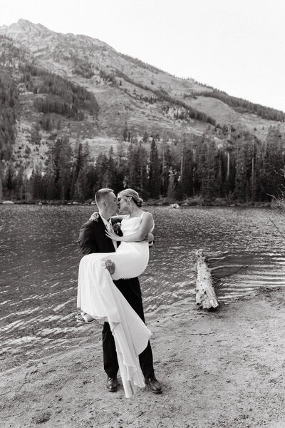 photo of groom holding bride next to a lake while they kiss with mountains in the background taken by Jackson Hole destination wedding photographer