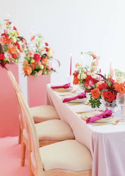 Elegant table setting with cream chairs, a light pink tablecloth, pink napkins, and pink and orange floral centerpieces