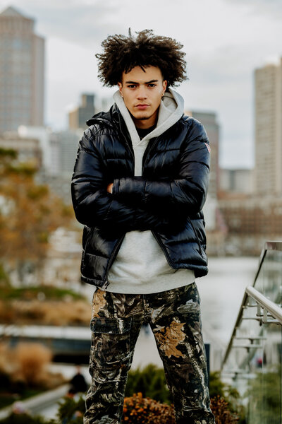 Boy wearing camo pants and a black puffy jacket standing with arms crossed in Boston.