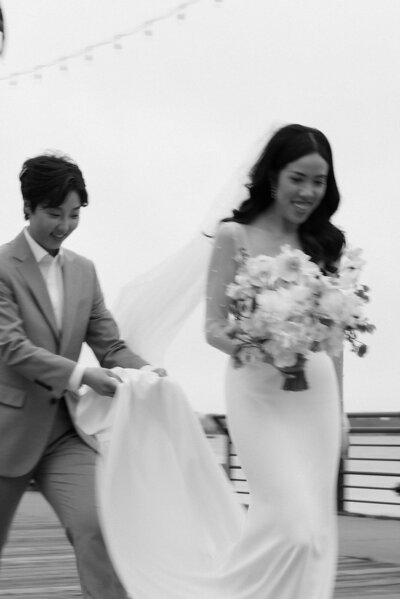 Groom holding bride’s train as she carries bouquet, black and white.