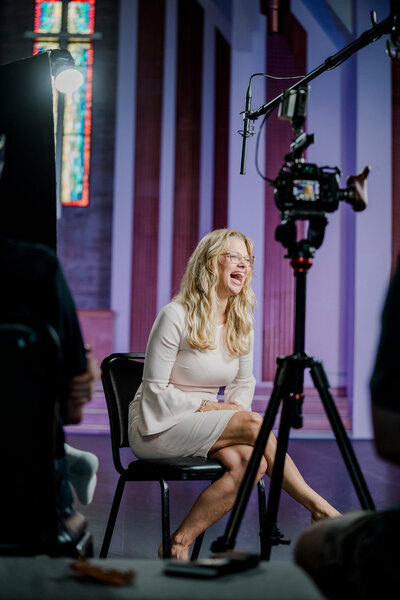 a woman laughs in front of a camera with a boom microphone hanging overhead