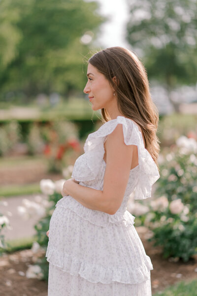 An expecting mom holding her belly while smelling a bush of pink flowers