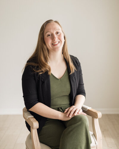 Strawberry blonde female, sitting in a chair, wearing a dark green pants and shirt with a black jacket over the green outfit, smiling at the camera
