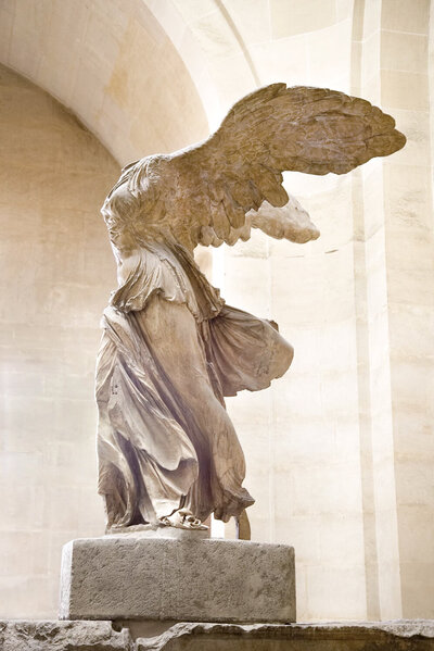 The image shows the Winged Victory of Samothrace, an ancient Greek statue of Nike, the goddess of victory. The statue is headless, with spread wings and draped clothing, standing on a stone base in a museum setting.