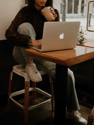 woman sits on a stool sipping her iced coffee while working on a laptop in a cafe