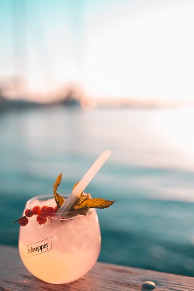 stemless cocktail glass with peach drink garnished with berries and greenery on a pier overlooking dark turquoise water