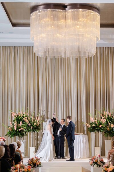 Bride and groom standing in front of a building with white pillars; the bride is holding a bouquet of red flowers