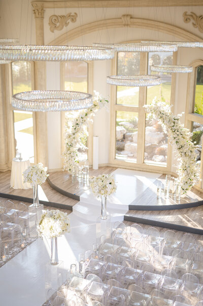 Stunning light filled wedding ceremony space with clear chairs, glass chandeliers, and white lush floral designs