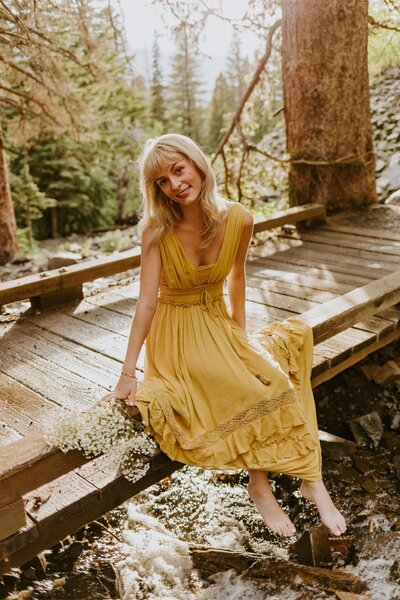Natalie sits on a bridge in a yellow dress. The lighting is very soft with lots of natural elements.