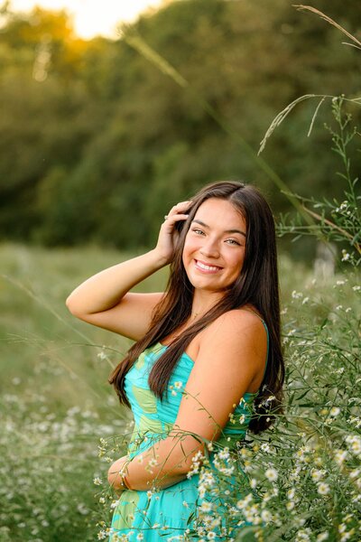 senior stands in a tall grass field surrounded by flowers with the sun falling on her hair
