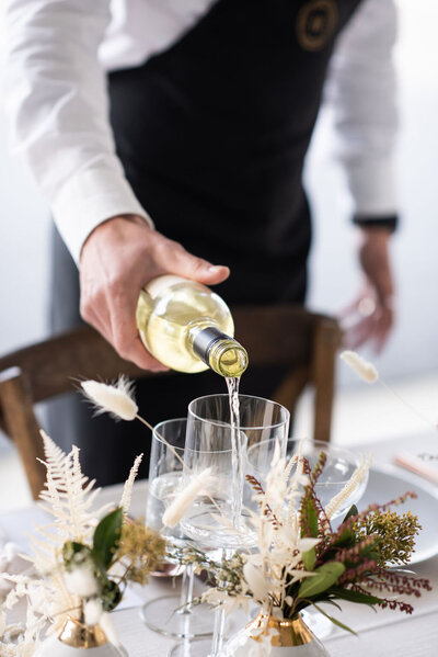 A well-dressed server pours a glass of white wine at table decorated for a wedding.