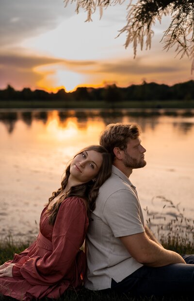 couple sitting back to back in front of a lake with the beautiful golden hour sunset behind them.