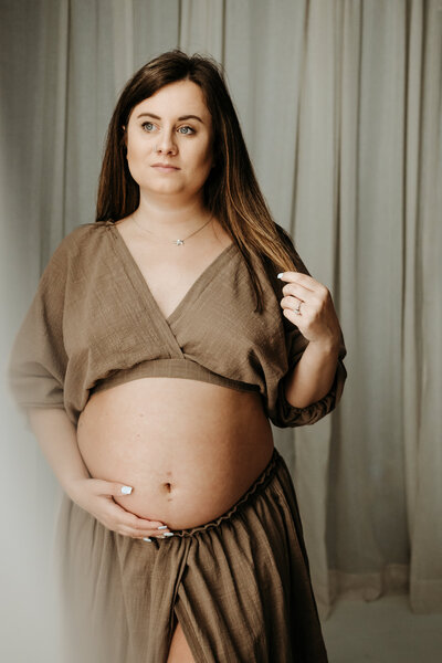 Maternity photo of pregnant mom twirling her hair and holding her belly in brown two piece dress in dark and moody studio in Maryland.