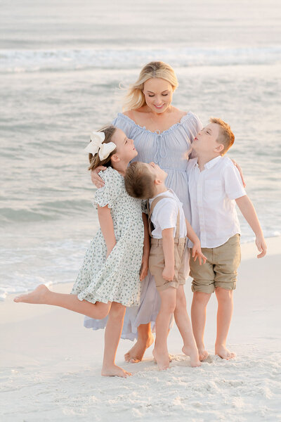 A graceful mother stands at the shoreline, smiling as her children snuggle close, framed by ocean waves and soft evening light.