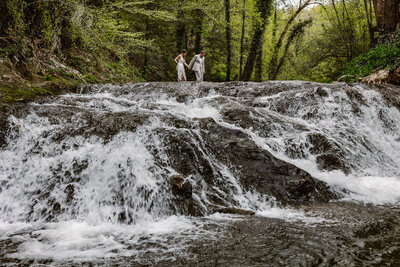 A couple walking across a waterfall on their elopement day. They are holding hands, and the groom is leading the way. 