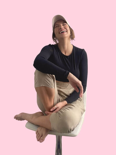 Caitlin sits comfortably on a chair, laughing with joy. She wears a black long-sleeve crop top, beige pants, and a light-colored cap against a pink background.