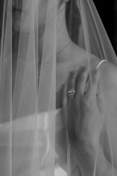 A bride and groom stand facing each other on a grassy lawn, captured perfectly by a skilled New England Wedding Photographer. The bride wears a long white gown with a flowing veil, holding a small bouquet. The groom is in a black suit, framed by vibrant pink flowers in the background.