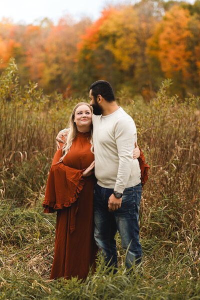 Couple looking at each other standing in a field during autumn with orange leaves on the tree in Ottawa, Ontario