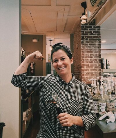 Woman smiles while holding a portafilter in one hand and flexing her arm 