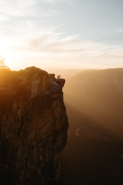 Couple stands on Taft Point cliff during their Yosemite Elopement with a gorgeous orange sunset behind them