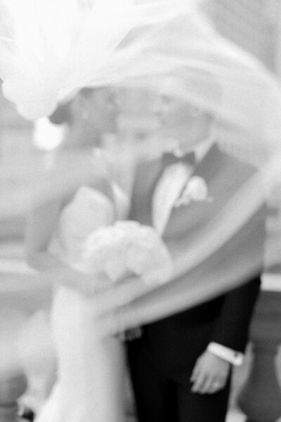 A romantic black and white wedding photo of a couple as the bride’s veil flows in front of the camera, creating a soft and cinematic effect.
