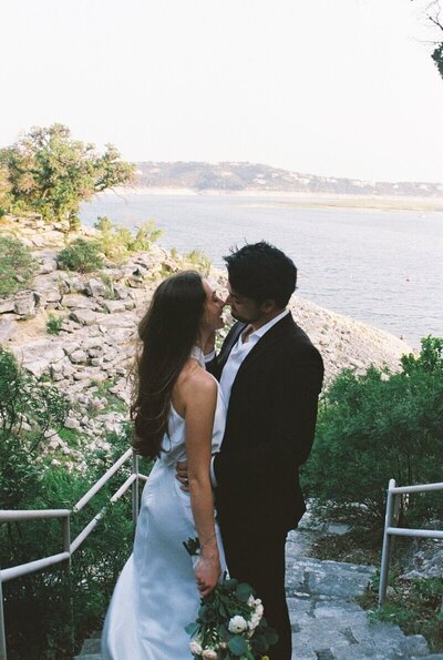 Austin elopement photographer captures candid moment between bride and groom on rocky lakeside staircase at Lake Travis, with scenic hill country view in the background.