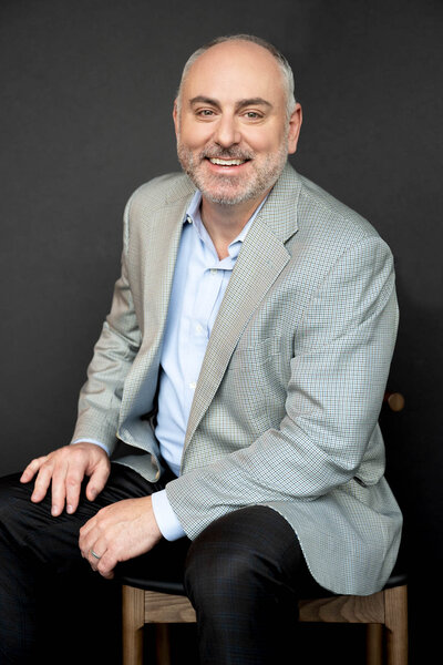 Headshot of a man wearing a grey jacket sitting on a stool smiling to the camera
