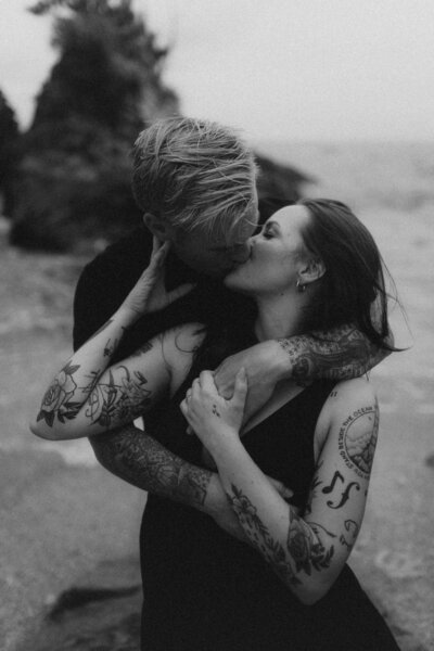 on the beach at sunset, A standing in front of J, arms wrapped around each other holding hands as J whispers into A's ear and gives her a kiss on her cheek